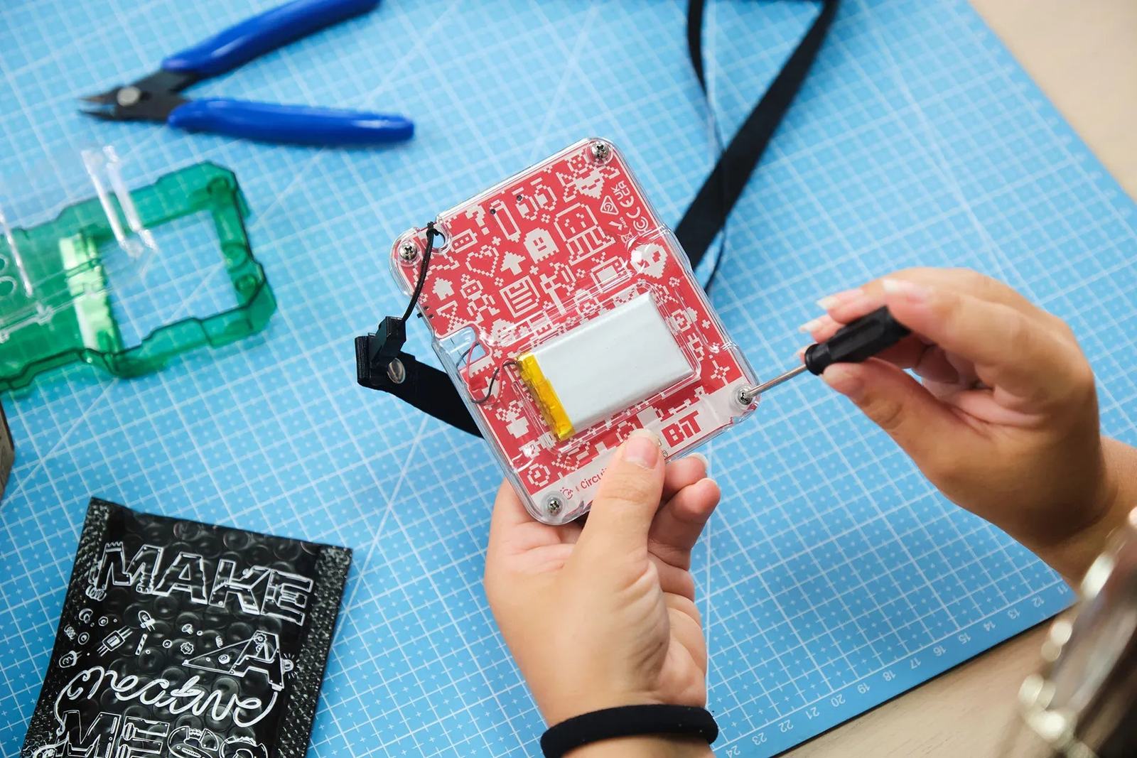 A child's hands holding a part-built CircuitMess Bit on a cutting mat, driving a screw into the clear case with the battery and red circuit board visible inside, side cutters and a spare case shell beside them.