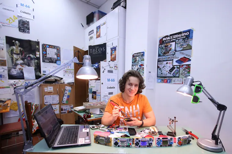 At the workshop desk in an orange tee, laptop open and half a dozen handheld consoles lined up under a lamp
