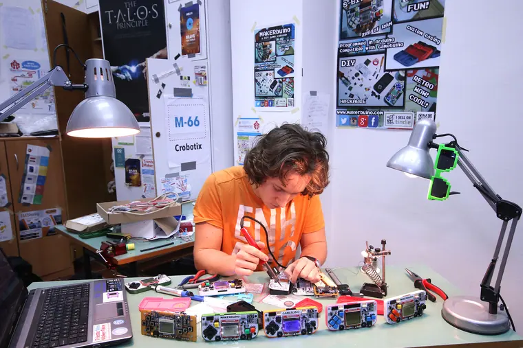Wide of the same bench session: laptop, desk lamps and a row of finished consoles laid out in front of him