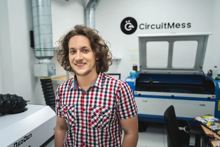 Standing in the CircuitMess workshop in a checked shirt, the laser cutter behind him and the company logo on the wall