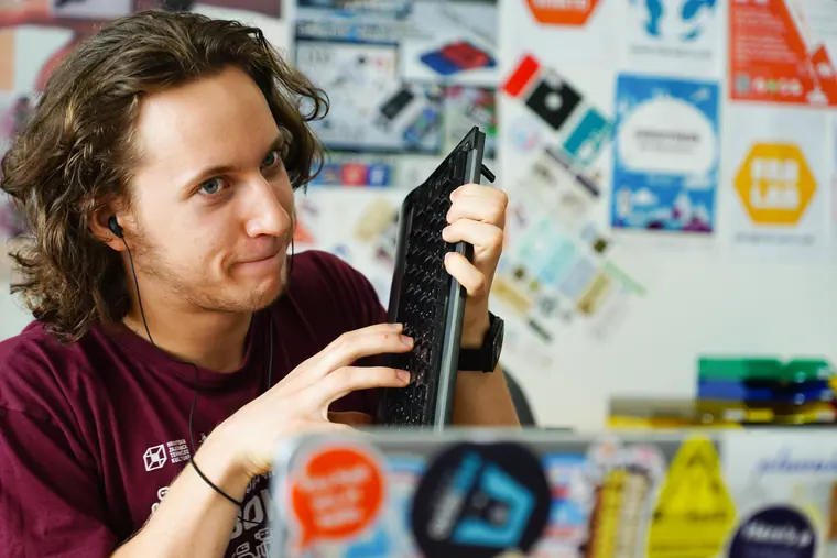 Candid at a desk, sticker-covered laptop open, lifting a bare board towards the light