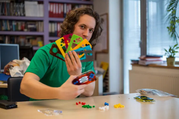 In a green tee, fanning out a spread of coloured console faceplates like a hand of cards, components scattered on the desk