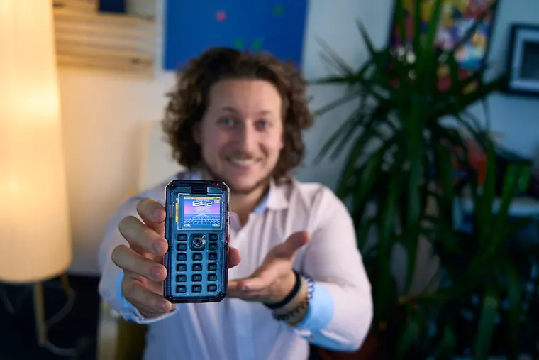 Holding a MAKERphone 2.0 out to camera with the screen lit, warm lamp behind
