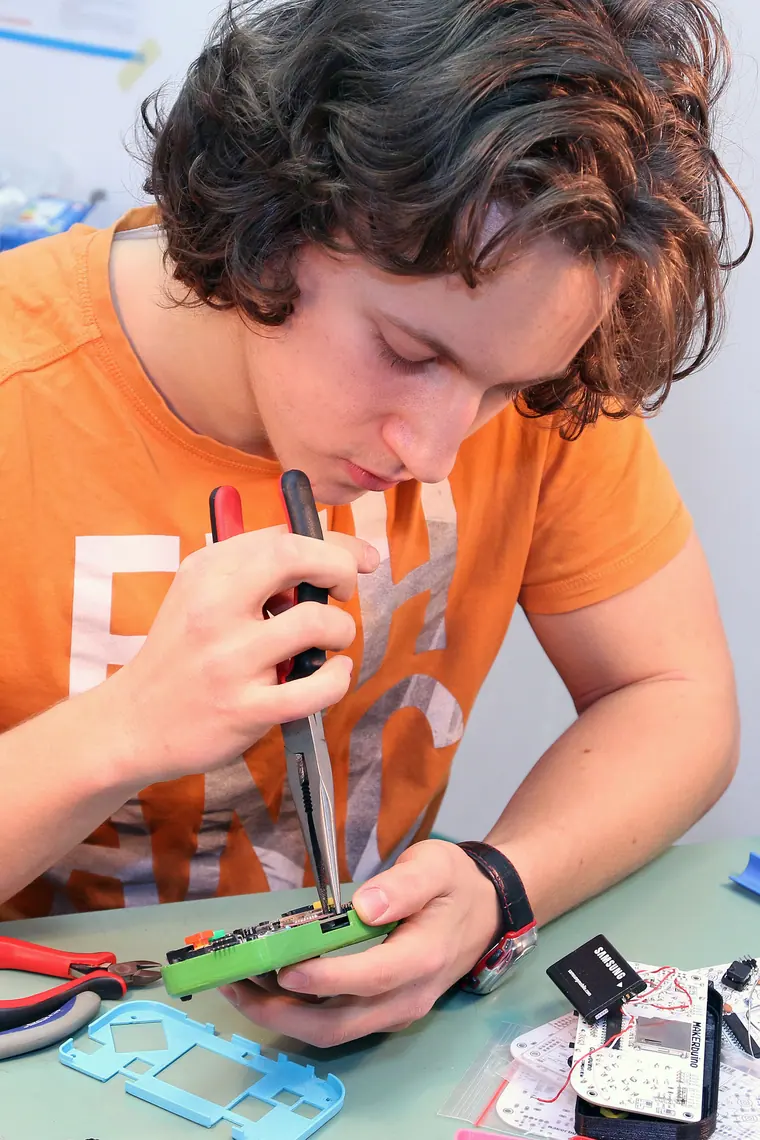 Soldering in an orange tee, tool in hand, leaning into a board on a green mat