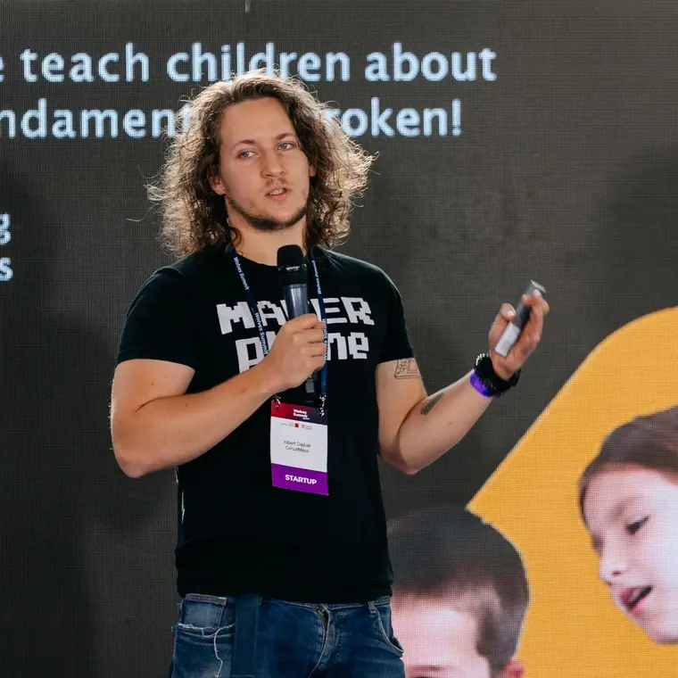 On stage with a hand mic and a lanyard, a child's face filling the screen behind him