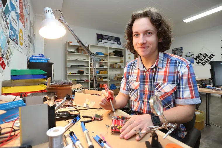 At the bench in a checked shirt under a desk lamp, hands on a part, workshop clutter and pinned notes filling the background