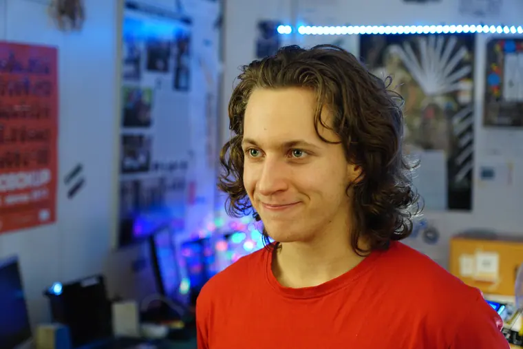 Head-and-shoulders in a red tee, turned to camera in the old workshop with shelving and boards out of focus behind